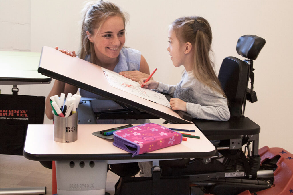 Child using a tiltable height adjustable desk with a therapist during a classroom activity 