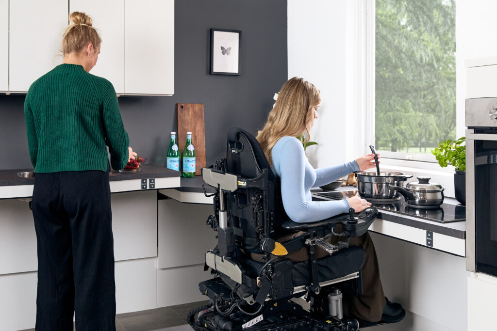 Two women cooking together in a Ropox accessible kitchen, one standing and one seated in a power wheelchair