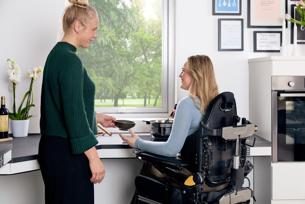 Wheelchair user smiling and handing a bowl to a standing person in a Ropox height-adjustable kitchen